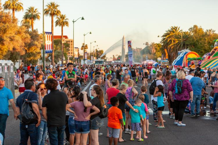 A crowd of people, mostly families, stand in a street surrounded by amusements and water slides in Mesa, AZ.